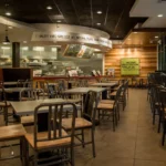 Modern Baja Fresh restaurant interior with neatly arranged tables and chairs, open kitchen in the background, and signage highlighting fresh, fire-grilled ingredients.